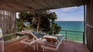 Una terraza de madera con sillones y una mesa, con vista a un mar tranquilo y abundante vegetación.