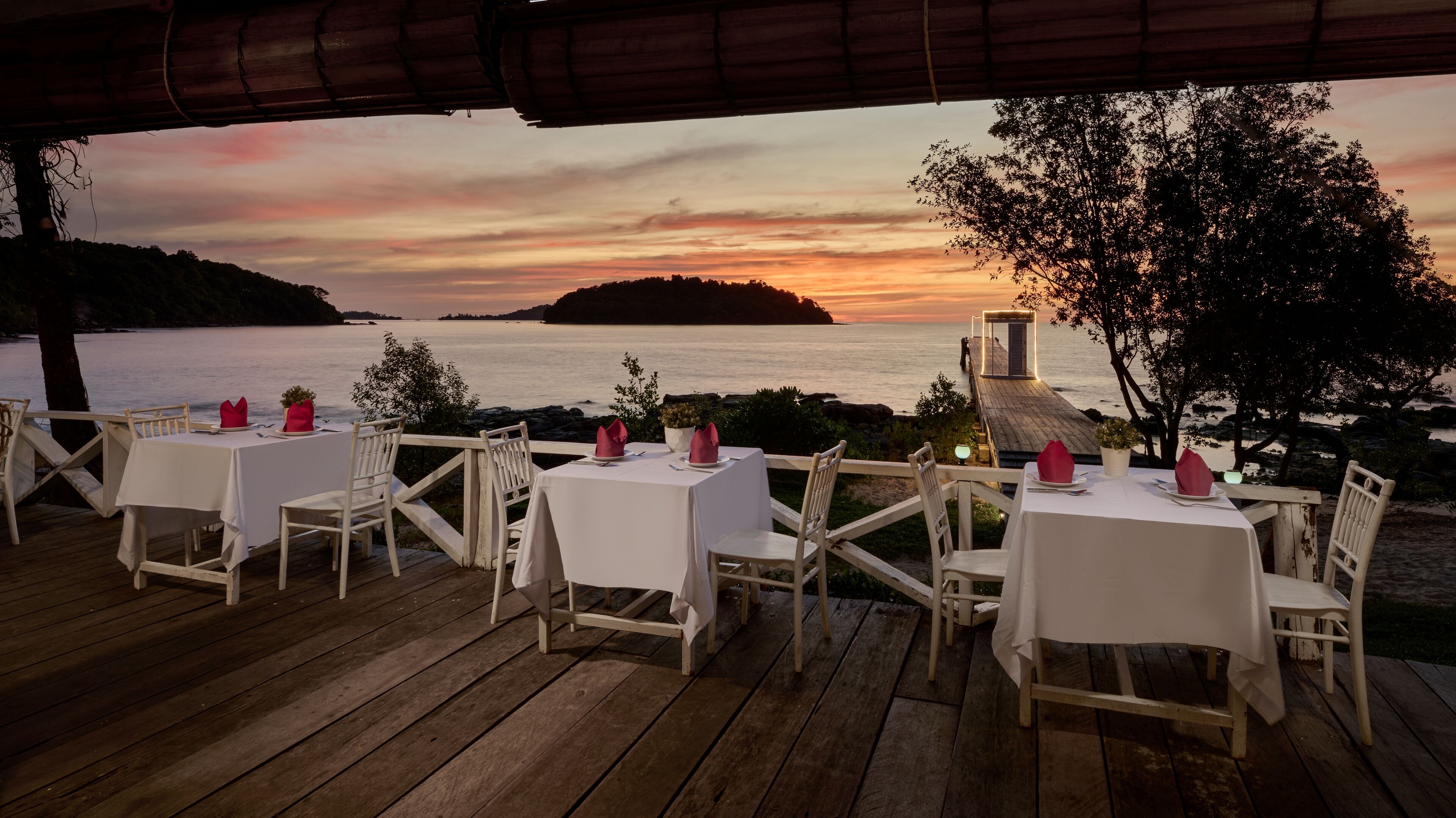 A dining area with tables and chairs set up on a wooden deck overlooking a body of water at sunset.