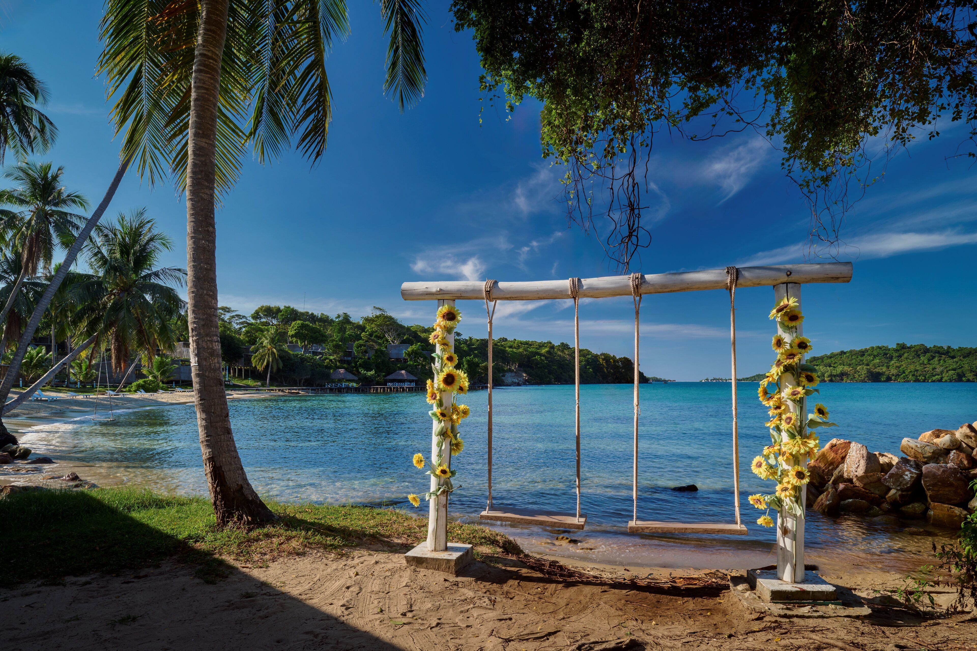 Hochzeitsdekoration am Strand mit einem Holzbogen, geschmückt mit Sonnenblumen, Palmen und freiem Blick auf das Meer.