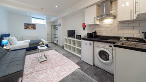 A modern hotel room with a bed, a gray sofa, a small table with a lamp, a washing machine, and a kitchenette.
