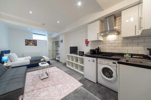 A modern hotel room with a bed, a gray sofa, a small table with a lamp, a washing machine, and a kitchenette.