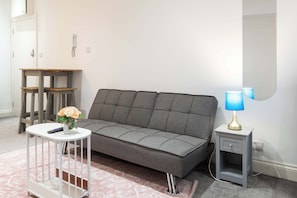 A modern living room with a grey sofa, a small white side table with a lamp, and a white side table with a vase of flowers. - Chic Shoreditch Studio,Fast Wi-Fi & Top Location (London)