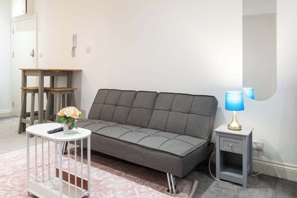 A modern living room with a grey sofa, a small white side table with a lamp, and a white side table with a vase of flowers.