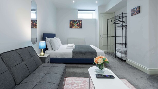 A modern bedroom with a bed, a sofa, a bedside table with a lamp, a small table with a vase of flowers, and a wall-mounted drying rack.