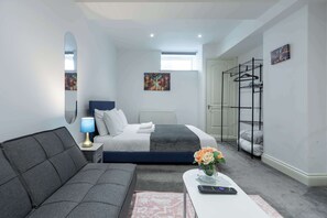 A modern bedroom with a bed, a sofa, a bedside table with a lamp, a small table with a vase of flowers, and a wall-mounted drying rack.