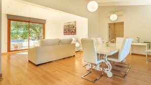 A modern living room with a white sofa, dining table, and chairs, large windows with a view of greenery, and a ceiling with exposed beams. - Villa Pancho - Large garden (Alcúdia)