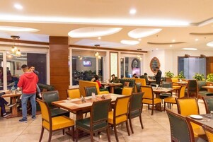 A restaurant with multiple dining tables and chairs, a TV screen in the background, and a few people dining.