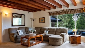 A living room with a sectional sofa, a wooden coffee table, a lamp, and a large window with a view of trees.