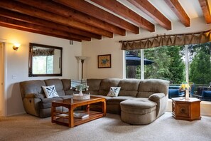 Una sala de estar con un sofá seccional, una mesa de centro de madera, una lámpara y un ventanal con vistas a árboles.
