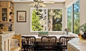 Un comedor con una mesa y sillas, un ventilador de techo y una ventana con vista a árboles.