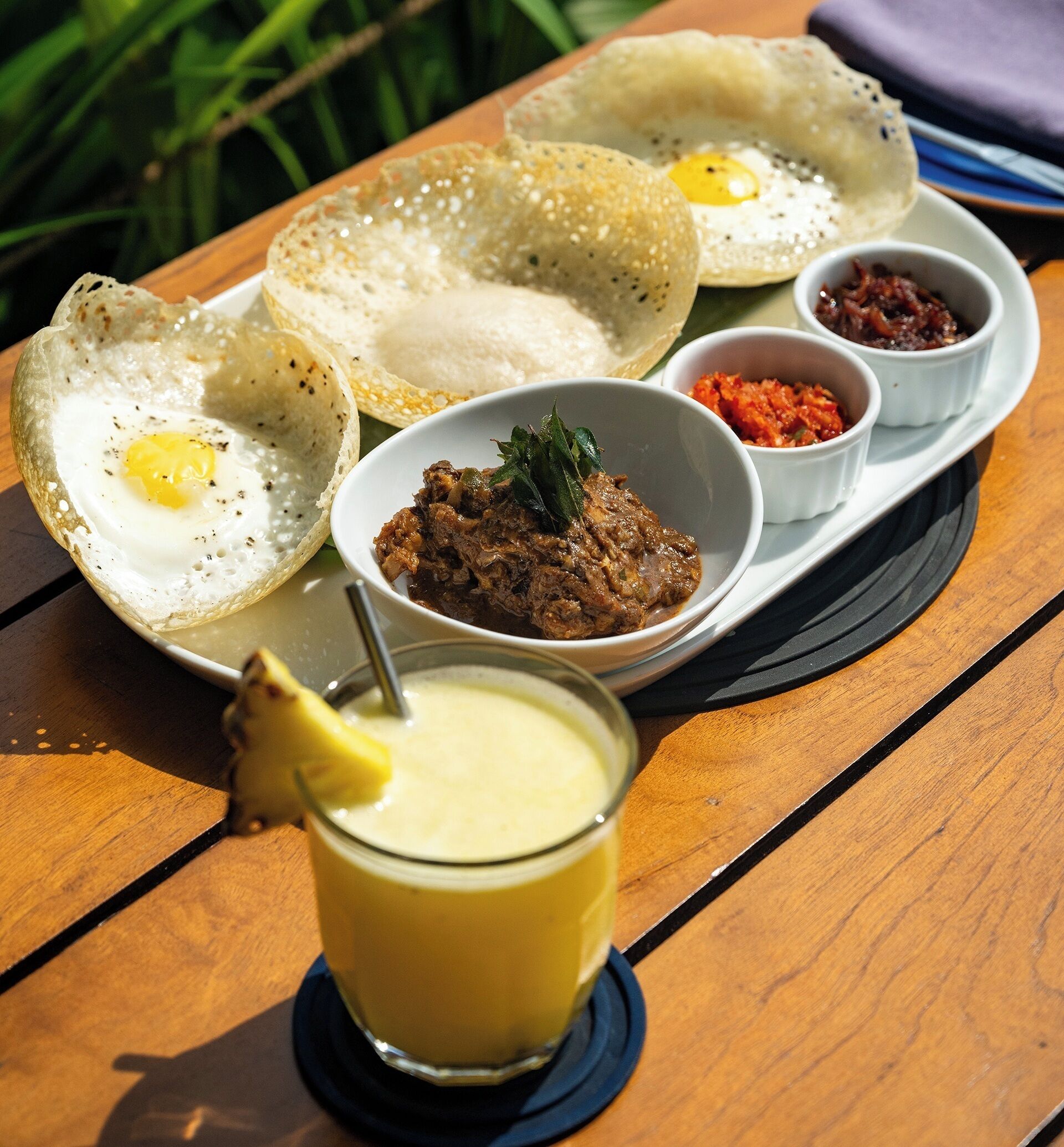 A meal with two crispy crepes, a bowl of meat dish, two small bowls of sauces, and a glass of yellow drink on a wooden table.