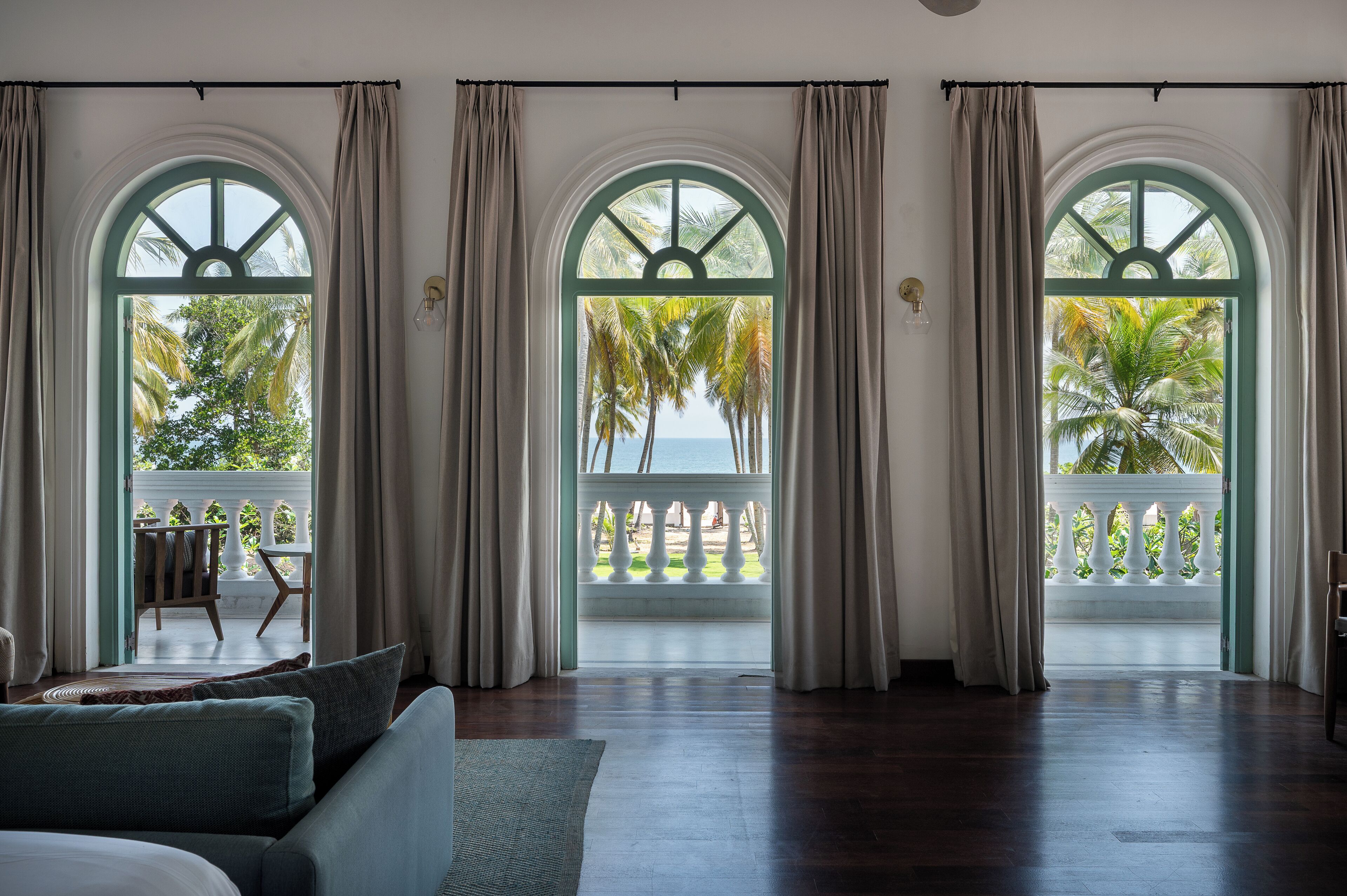 Una habitación con un amplio ventanal arqueado, un balcón con barandilla blanca y vistas a palmeras y al mar.