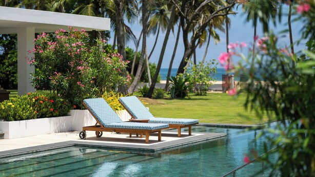 A pool area with two lounge chairs, surrounded by tropical plants and palm trees, with a clear view of the ocean. - Kalkudah Beach House (EP)