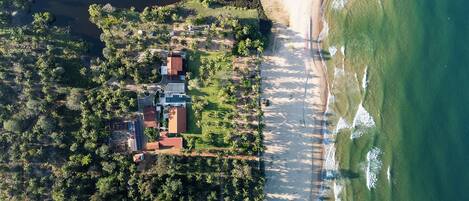 An aerial view of a coastal property with a beach, dense vegetation, and a few buildings.