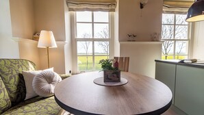 A cozy room with a round wooden table, a green patterned sofa, and a view of a green field through large windows. - Lüsthuus vacation home - historic charm meets modern comfort (Krummhörn)