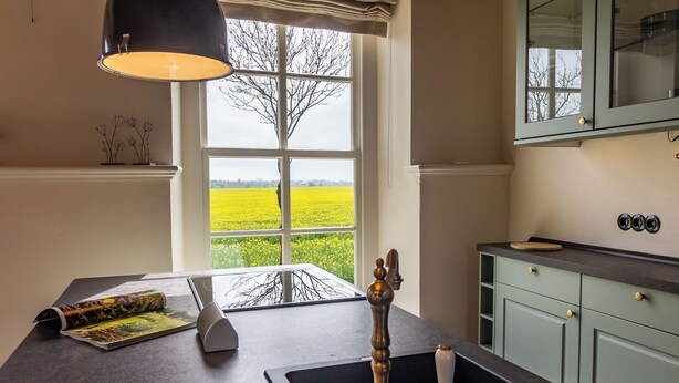 A kitchen with a window view of a yellow field, a sink with a vintage faucet, and a cabinet with a light green door. - Lüsthuus vacation home - historic charm meets modern comfort (Krummhörn)