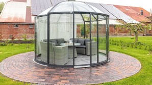 A glass-enclosed gazebo with wicker furniture and a brick circular patio. - Lüsthuus vacation home - historic charm meets modern comfort (Krummhörn)