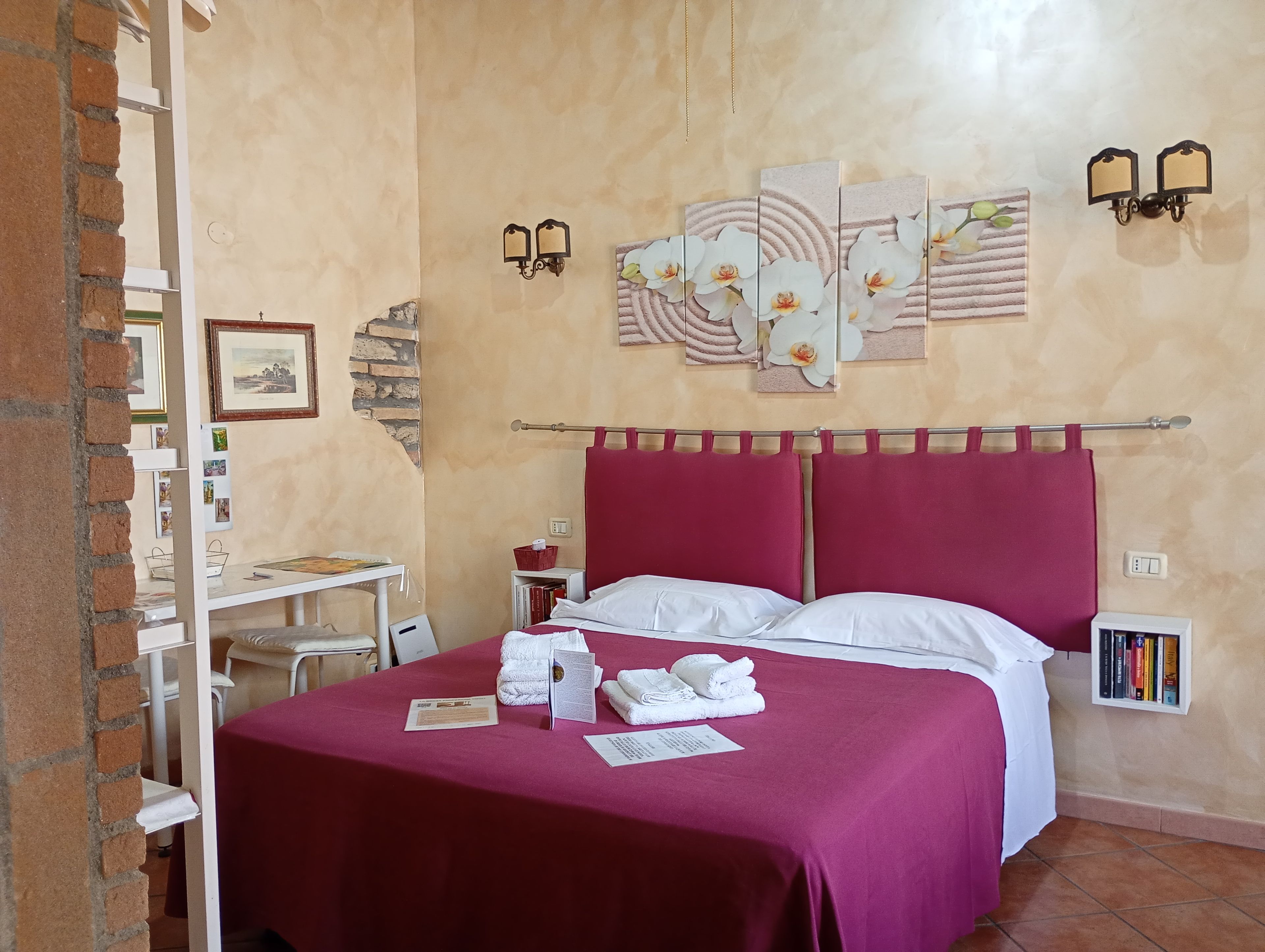 A hotel room with a bed, a small table, a shelf with books, and a wall with artwork.
