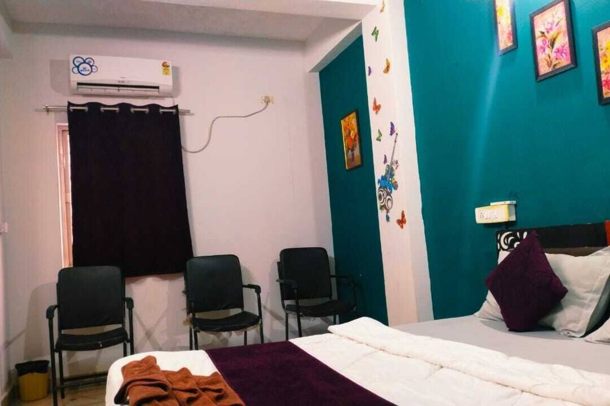 Ένα δωμάτιο με ένα κρεβάτι, δύο καρέκλες, μια  κουρτίνα, μια μονάδα κλιματισμού και έναν τοίχο διακοσμημένο με σχέδια λουλουδιών.