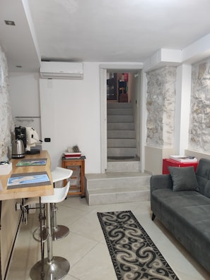 Una sala de estar moderna con un sofá gris, una zona de cocina con un taburete de bar y una escalera que conduce a un nivel superior.