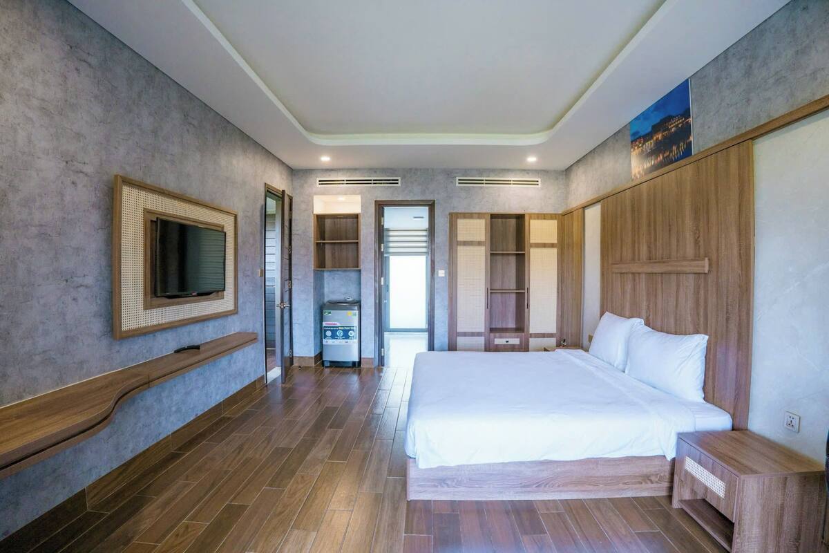 A modern hotel room with a large bed, built-in wardrobe, a television mounted on the wall, and a trash bin.