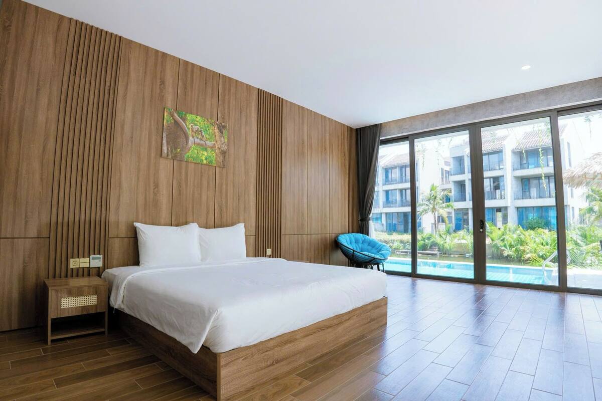 A modern hotel room with a large bed, wooden panel walls, a small side table, and a view of a swimming pool and greenery outside.