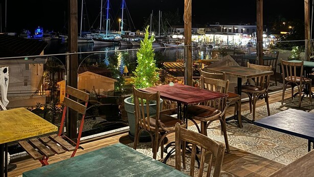 Ein Essbereich im Freien mit Holztischen und -stühlen, ein kleiner Weihnachtsbaum und Blick auf Boote und einen Yachthafen bei Nacht.