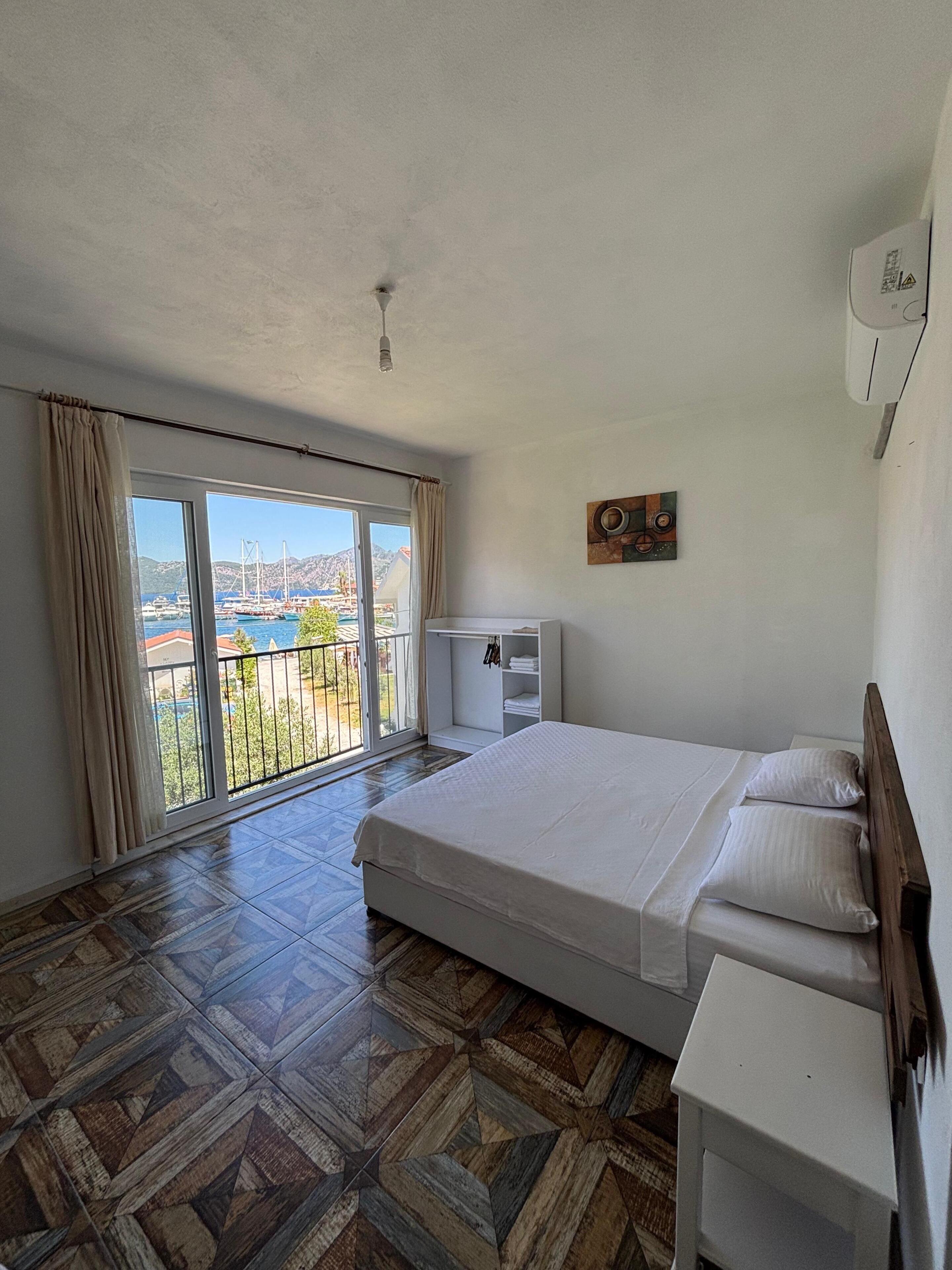 A bedroom with a large bed, a small bedside table, a wardrobe, and a view of a marina through a large window.