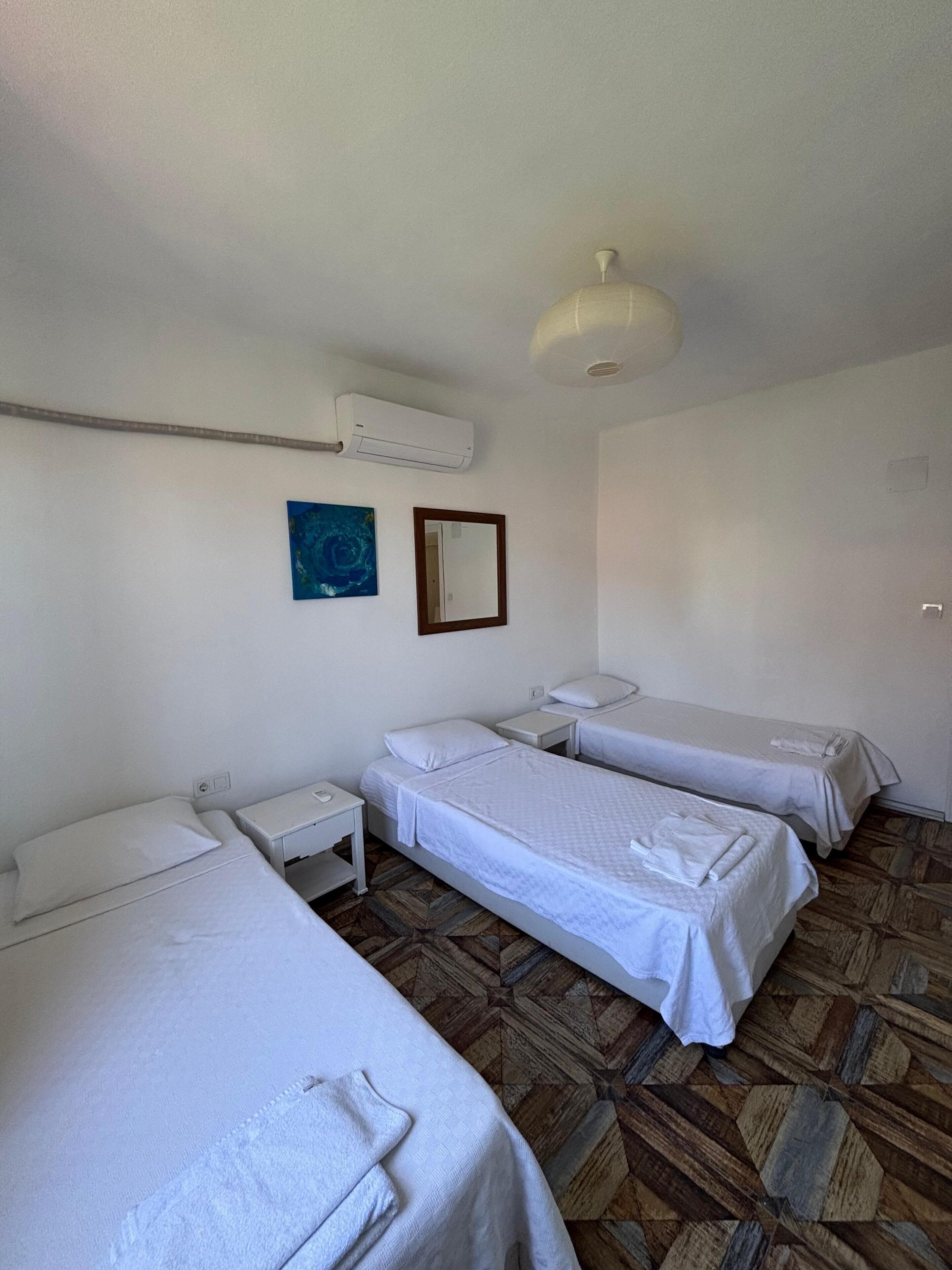 Habitación de hotel con tres camas individuales, una mesita, aire acondicionado instalado en la pared, un espejo y un cuadro azul.