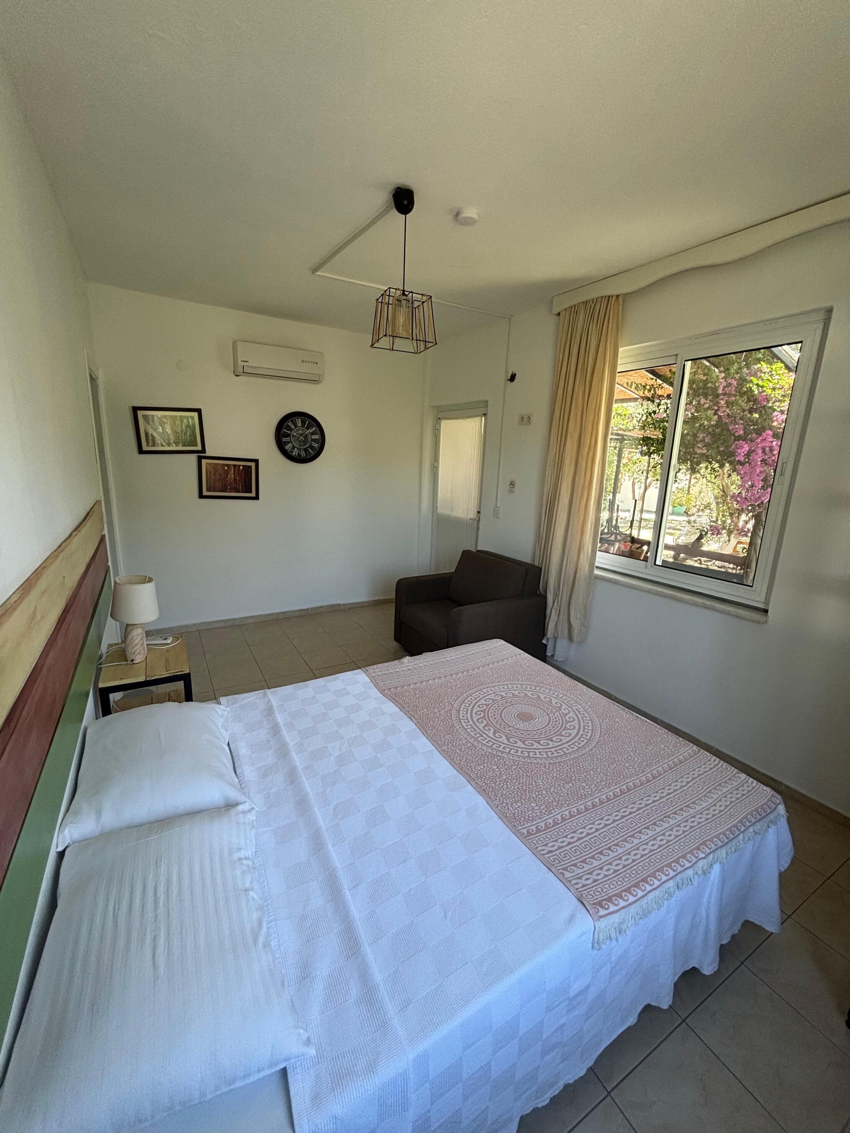 A bedroom with a bed, a nightstand, a lamp, a ceiling fan, a window with curtains, and a wall clock.