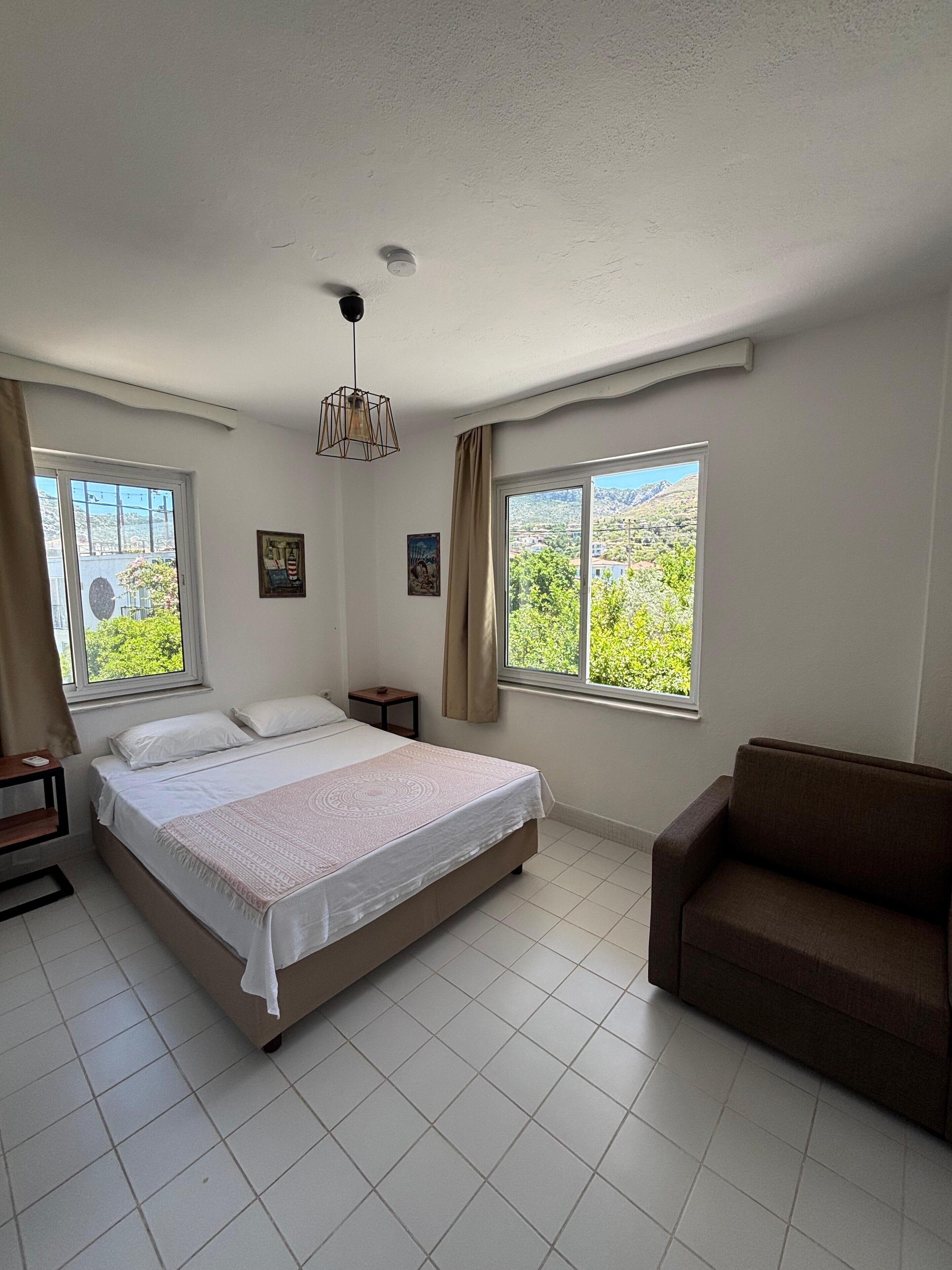 A bedroom with a bed, a sofa, and two windows offering a view of greenery and buildings.