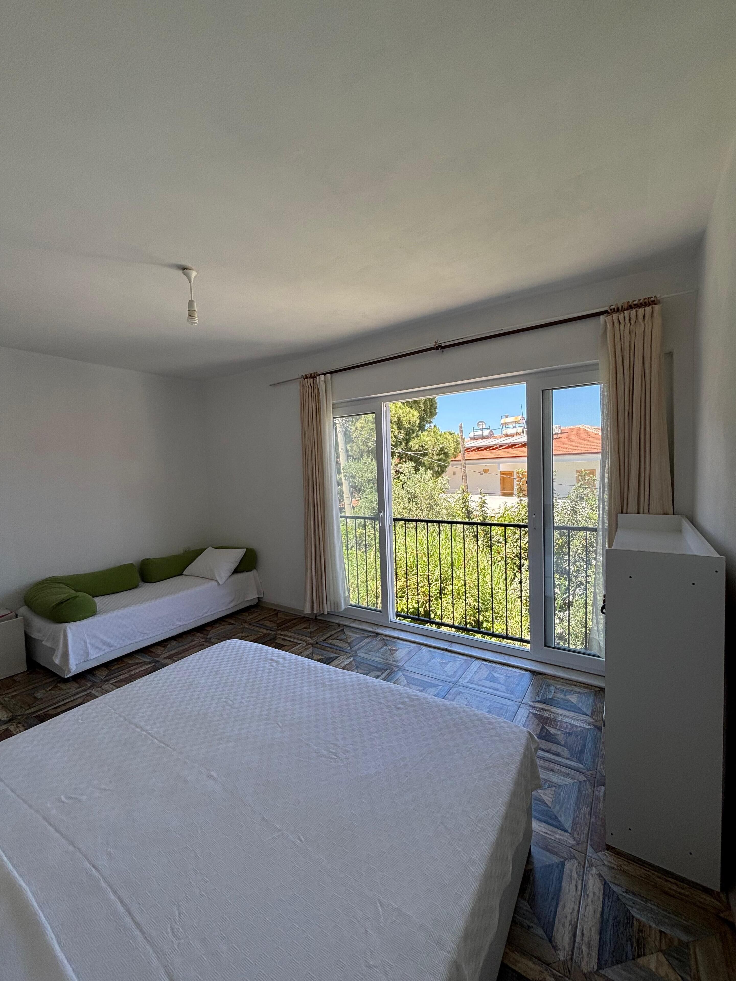 A bedroom with a bed, a sofa, a window with a view of the outside, and a ceiling fan.