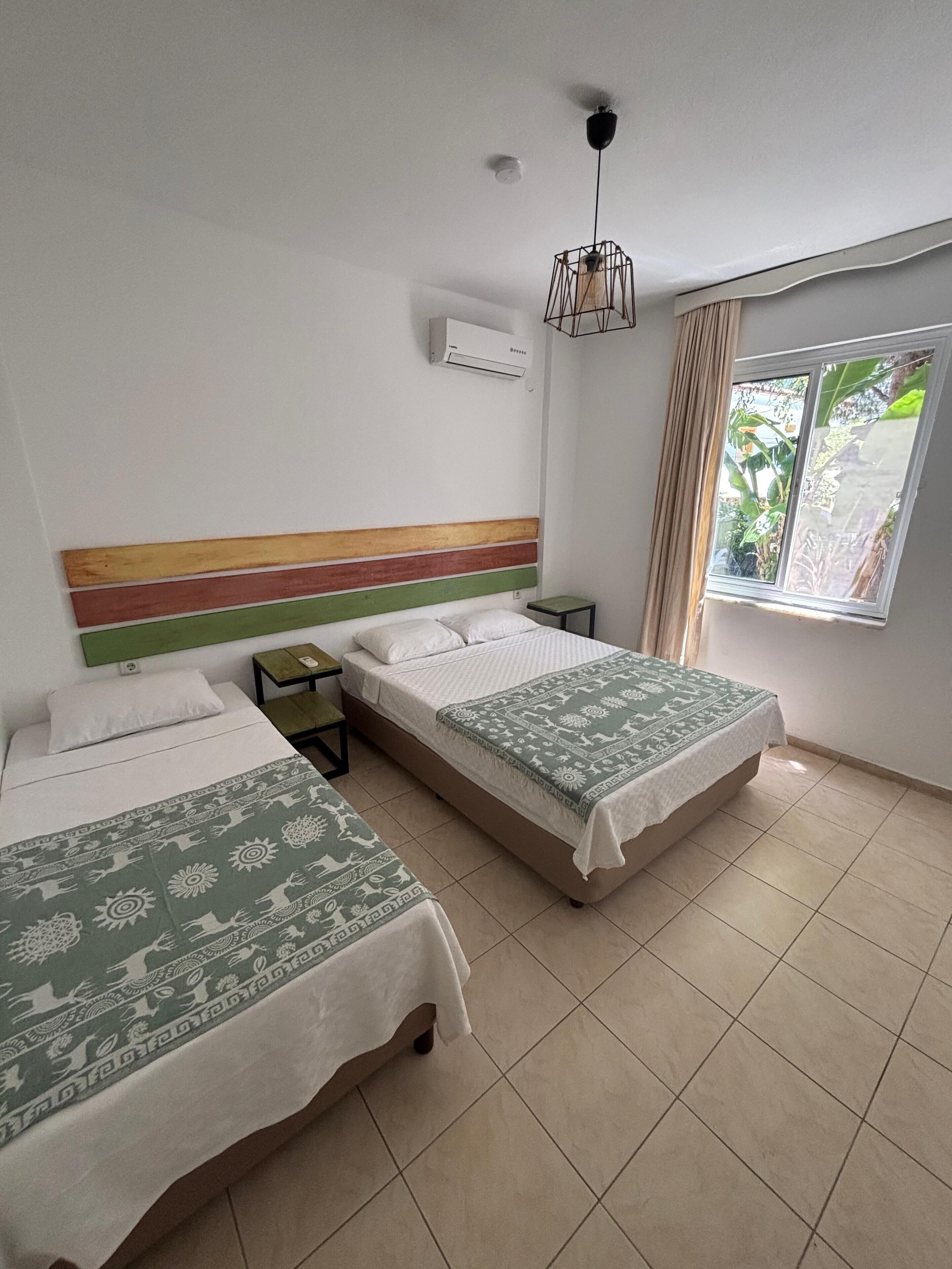 A room with two beds, a window with curtains, a hanging light fixture, and an air conditioning unit.