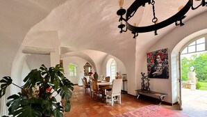 Un comedor amplio con una planta grande, un candelabro y vistas al jardín a través de ventanas arqueadas.