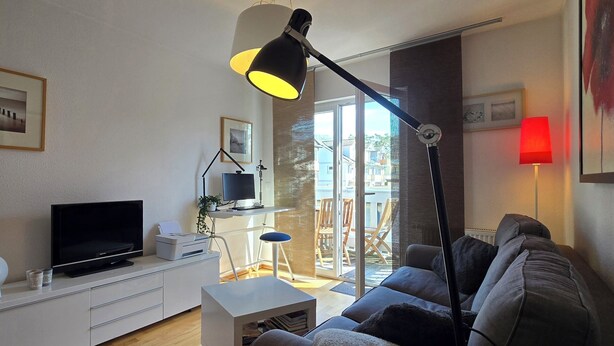 Un salon moderne avec un téléviseur à écran plat, un meuble blanc, un lampadaire noir, un canapé gris et une lampe rouge.