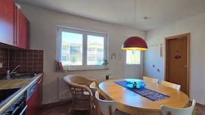 Une cuisine compacte avec un coin repas, comprenant une table ronde, des chaises en osier et un luminaire suspendu rouge.