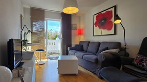 Un salon moderne avec un canapé gris, une table basse blanche et un fauteuil noir. Une grande fenêtre offre une vue sur un balcon et des arbres.