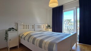 Une chambre à coucher avec un lit, des tables de chevet, une petite table, une plante en pot et une vue sur des immeubles par la fenêtre.