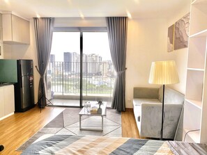 A modern hotel room with a city view, a kitchenette, a sofa, a lamp, and a coffee table. - Radiant Vinhomes Smart City - Apartment (Hanoi)