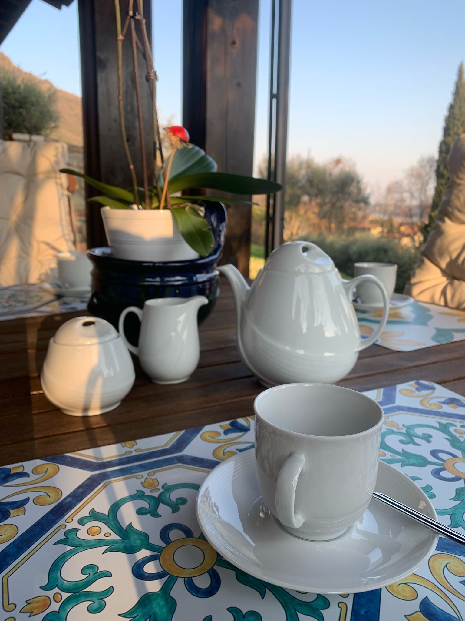 A table set with white teacups and a teapot on a patterned tablecloth.