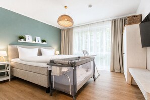 A modern bedroom with a large bed, a baby crib, a bedside table, a lamp, a wall-mounted shelf with decorative items, and a window with curtains.