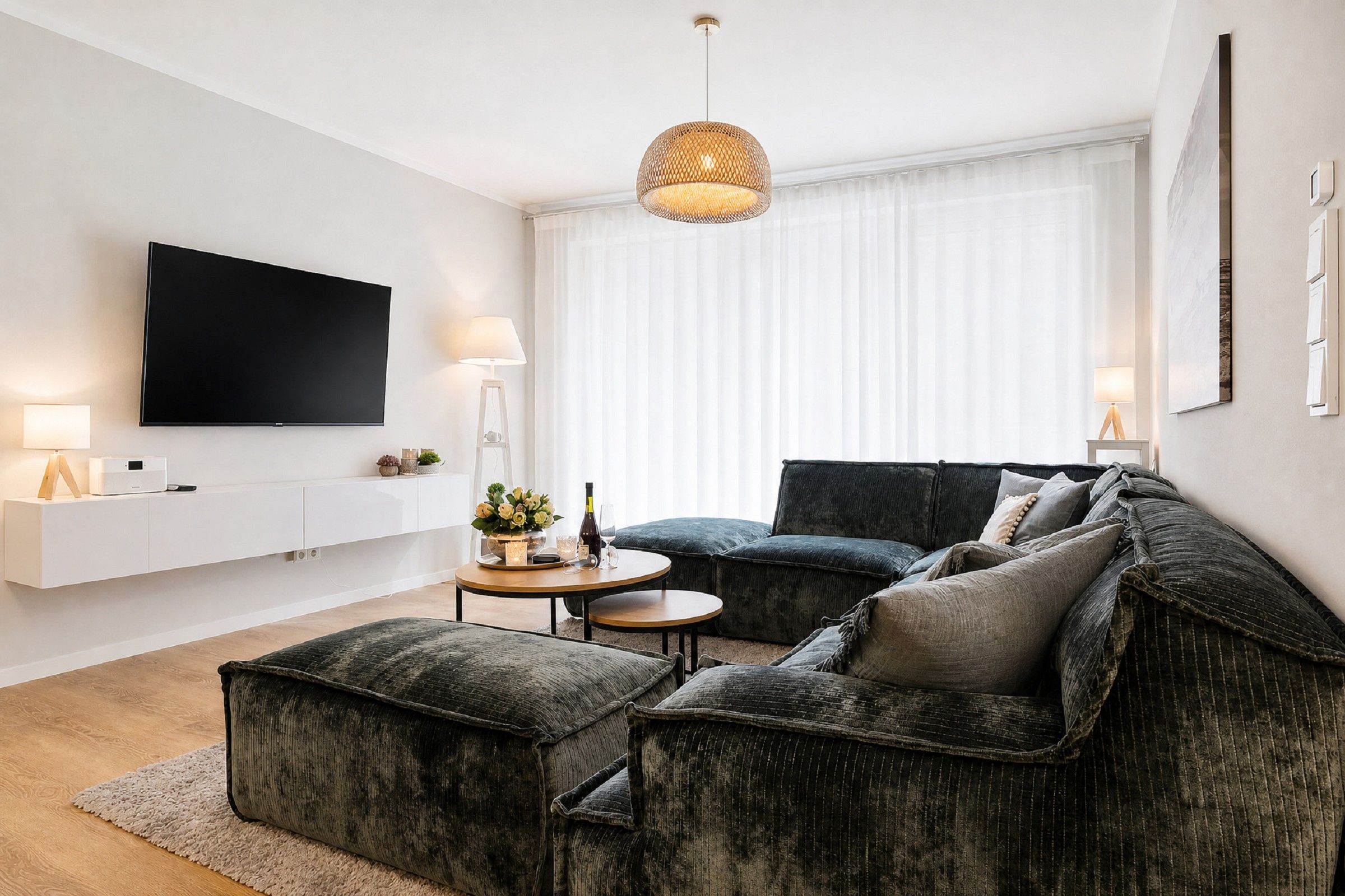 A modern living room with a flat-screen TV, a round coffee table, a dark sofa, and a large window with sheer curtains.