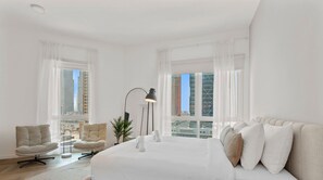 A modern bedroom with a large bed, two armchairs, a floor lamp, and large windows with city views.