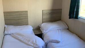 A small, compact room with two single beds, a wooden headboard, and a window with curtains. - Seaside Retreat - Golspie Cabin (Golspie)