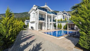 A large, modern house with a swimming pool, multiple balconies, and a mountain backdrop.