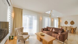 A modern living room with a brown leather sofa, a beige armchair, a wooden coffee table, and a dining area with a table and chairs.
