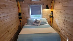 Una stanza in legno, stretta, con un letto, una finestra con persiane e due tavole da surf appoggiate alle pareti.