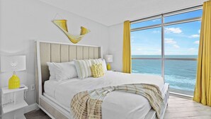 Un dormitorio con una cama grande, vista al océano y una ventana con cortinas amarillas.
