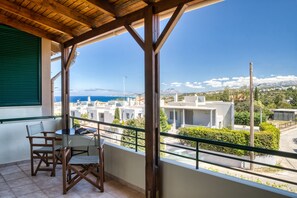 Un balcón con vistas a un pueblo costero, muebles de madera y una ventana con persiana verde.