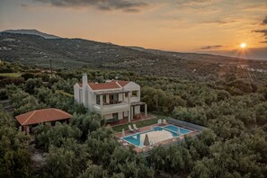 A modern villa with a swimming pool, surrounded by dense vegetation and hills in the background.